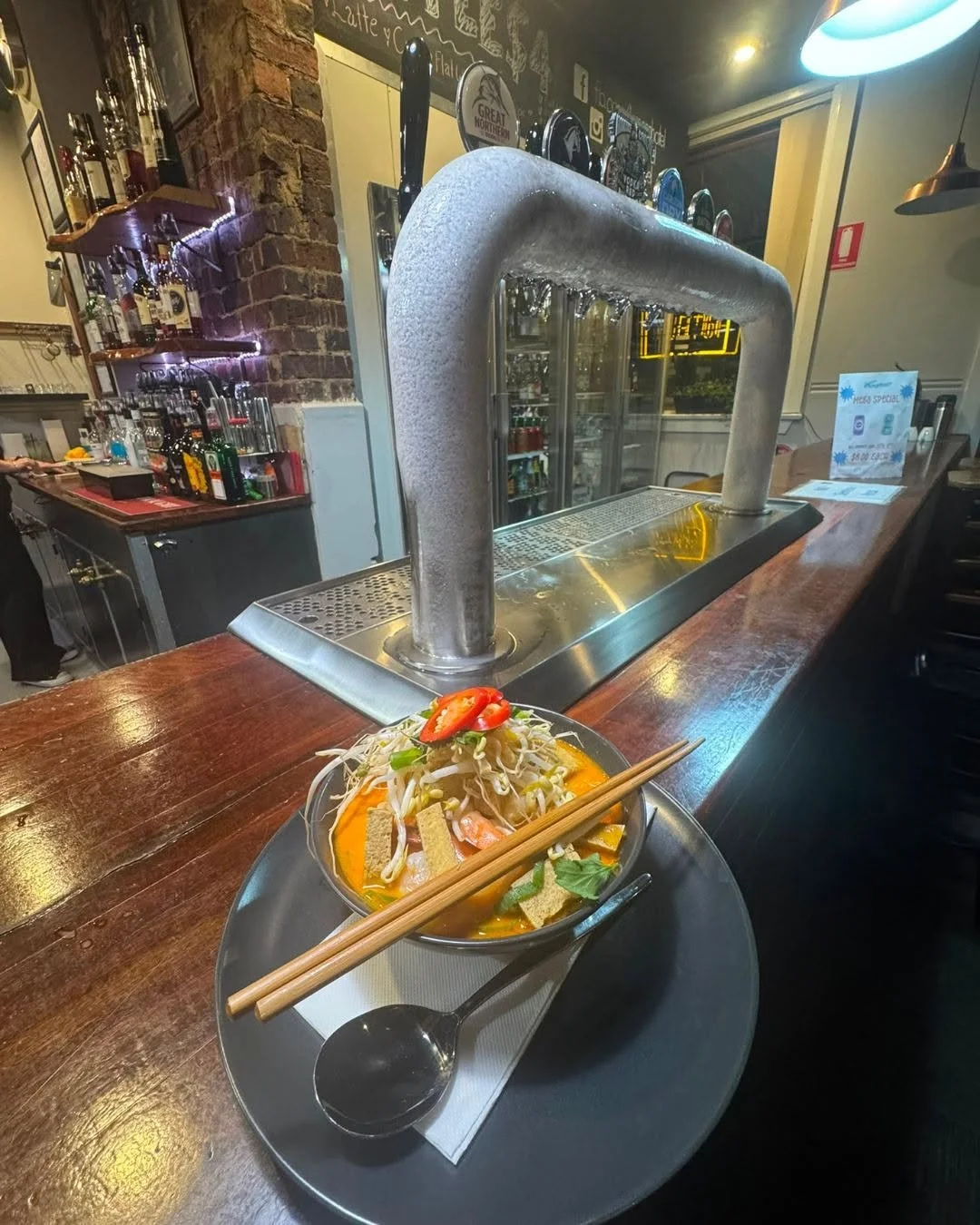 A bowl of Asian noodle soup with vegetables and sliced chili peppers on top, placed on a black tray with wooden chopsticks and a spoon, on a wooden bar counter in a restaurant.