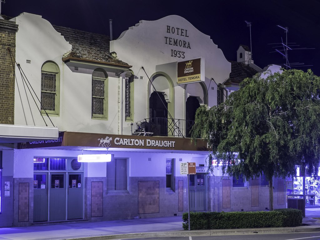 Nighttime view of a historic two-story building with a sign saying 'Hotel Temora 1933' and advertisements for Carlton Draught beer. The building has arched windows, some with stained glass, and a small balcony. A large leafy tree partially obscures t