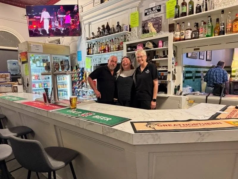 Three people smiling behind a bar with shelves of alcohol bottles, a TV screen showing a music performance, and chalkboard signs, in a pub or bar setting.