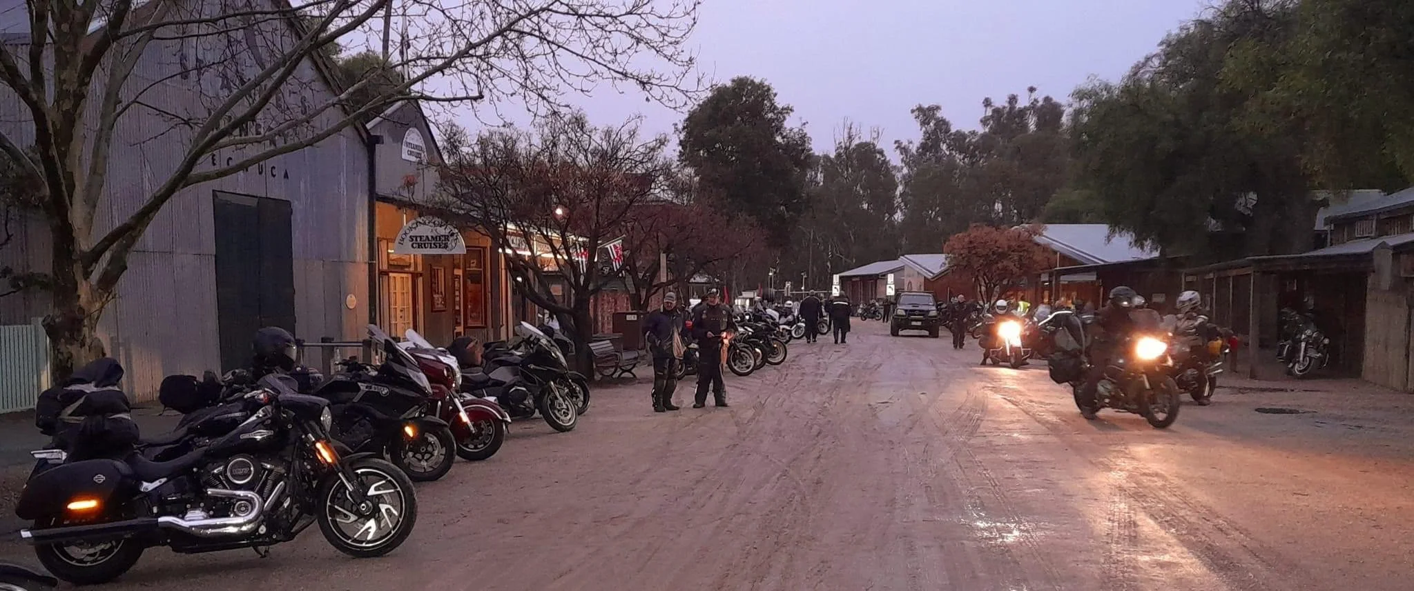 A dirt street with parked motorcycles on the left and people walking or standing around, some wearing helmets. Buildings on both sides of the street include a shop with a sign that says 'Steamers Cruise'. The sky appears to be at dusk or dawn, with t