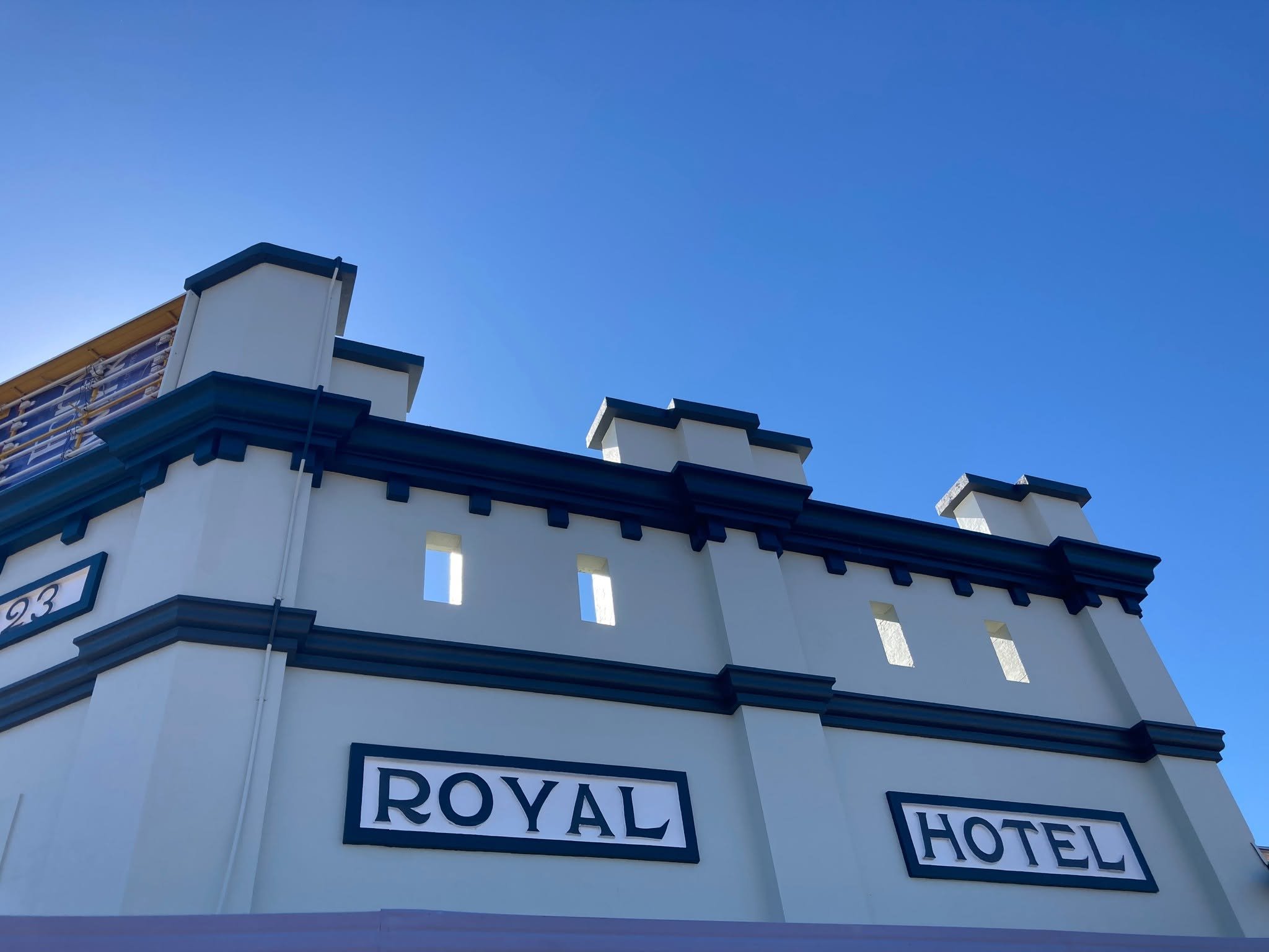 The image shows the upper part of a white hotel building with black and white architectural details, a sign reading 'Royal Hotel,' and a clear blue sky in the background.