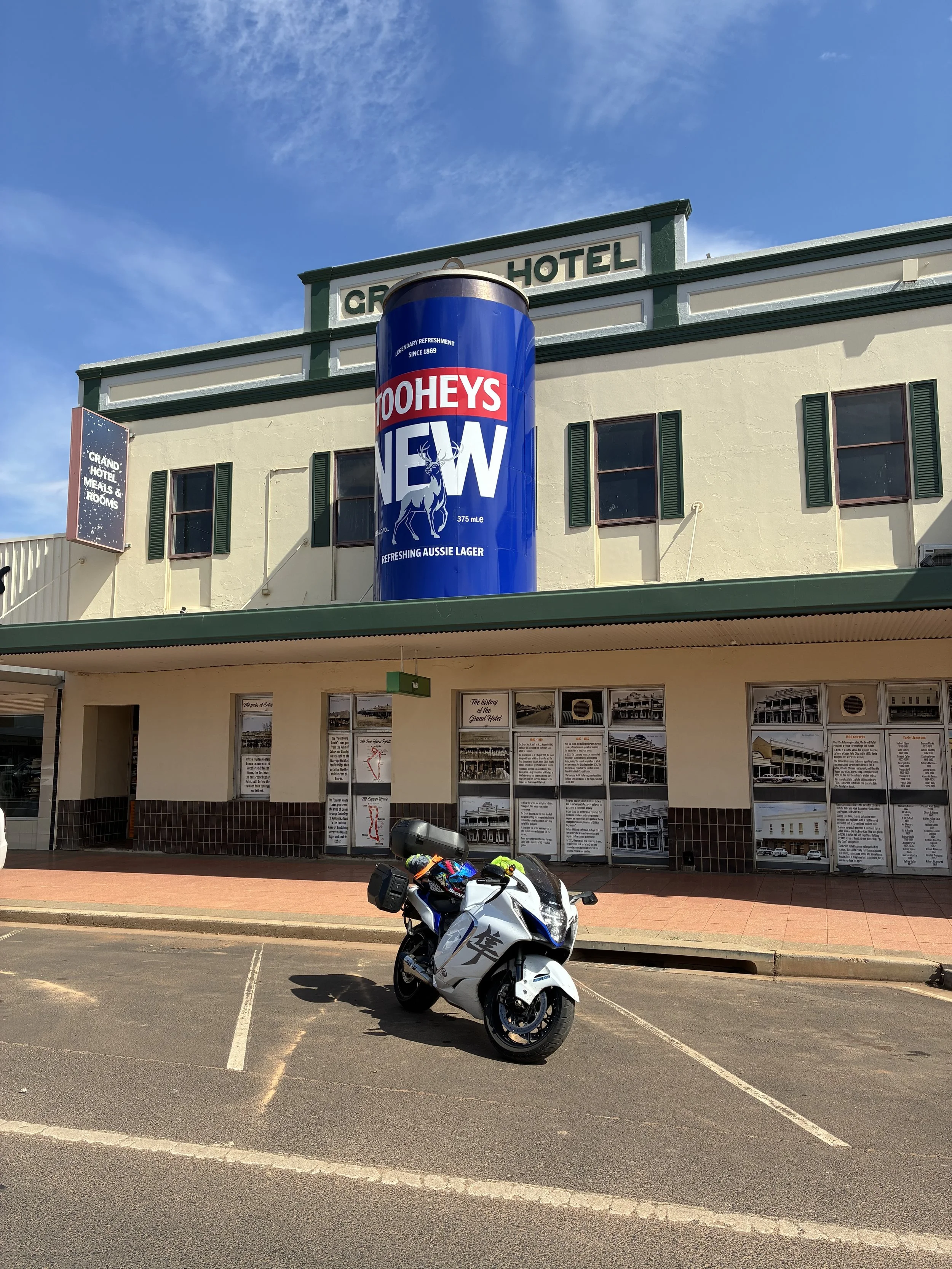 A white motorcycle parked in a parking lot in front of a building with historical photos and information, with a large blue can of Tooheys beer on top of the building and the word "HOTEL" visible at the top.