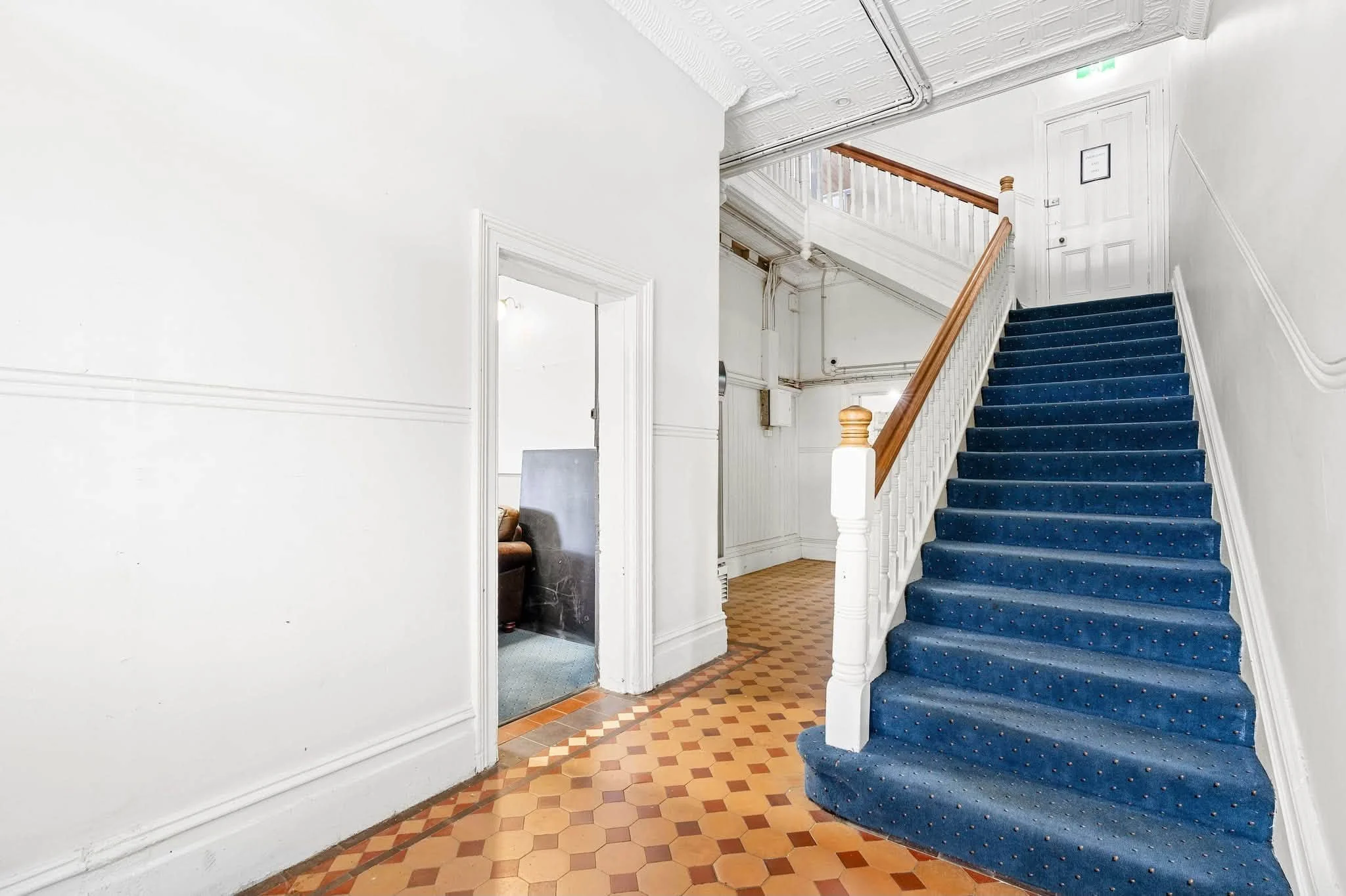 Interior of a house with a staircase covered by a blue carpet, white walls, and a hallway with patterned tile floor.
