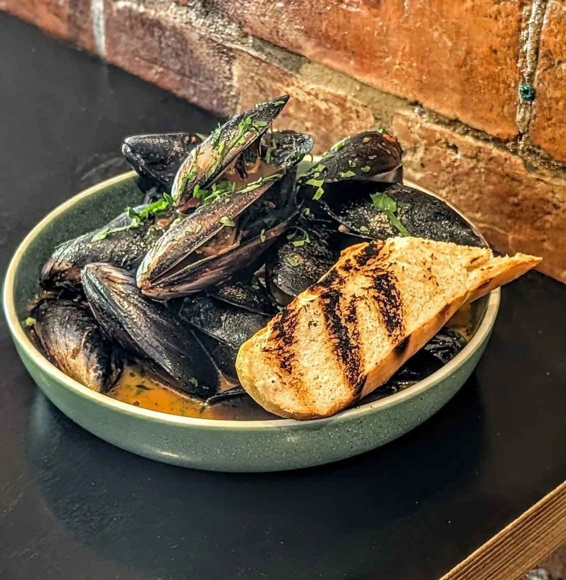 A bowl of cooked mussels garnished with green herbs, served with toasted bread in the foreground on a black surface, with a brick wall in the background.
