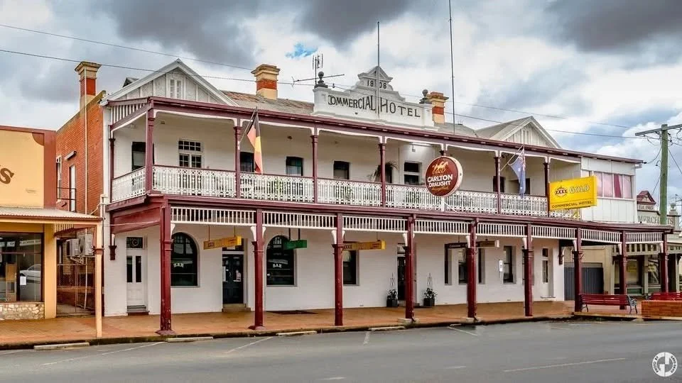 Two-story white and red building labeled 'Commercial Hotel' with a balcony on the second floor, hanging signs for Carlton Draught beer and XXX Gold, and a cloudy sky in the background.