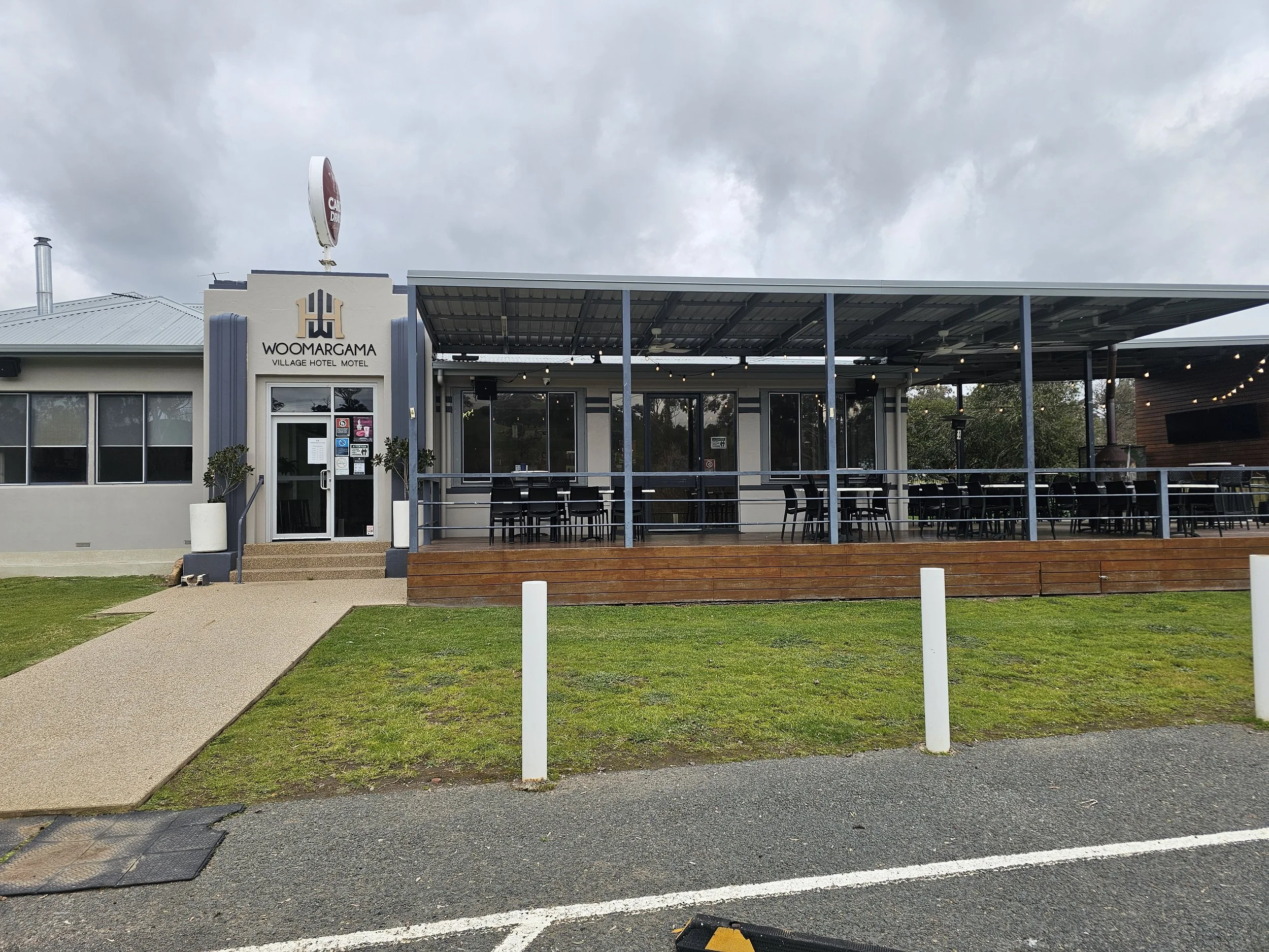 Exterior view of Woomargama Village Hotel Motel with a covered outdoor seating area, entrance door, and signage, under a cloudy sky.