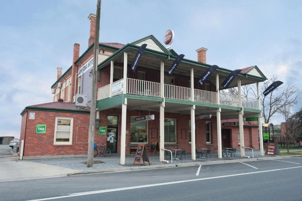 A two-story brick building with a covered porch on both levels, featuring seating outside and numerous black flags hanging from the upper balcony. The building appears to be a hotel or guesthouse, with signs indicating lodging and a shop on the premi