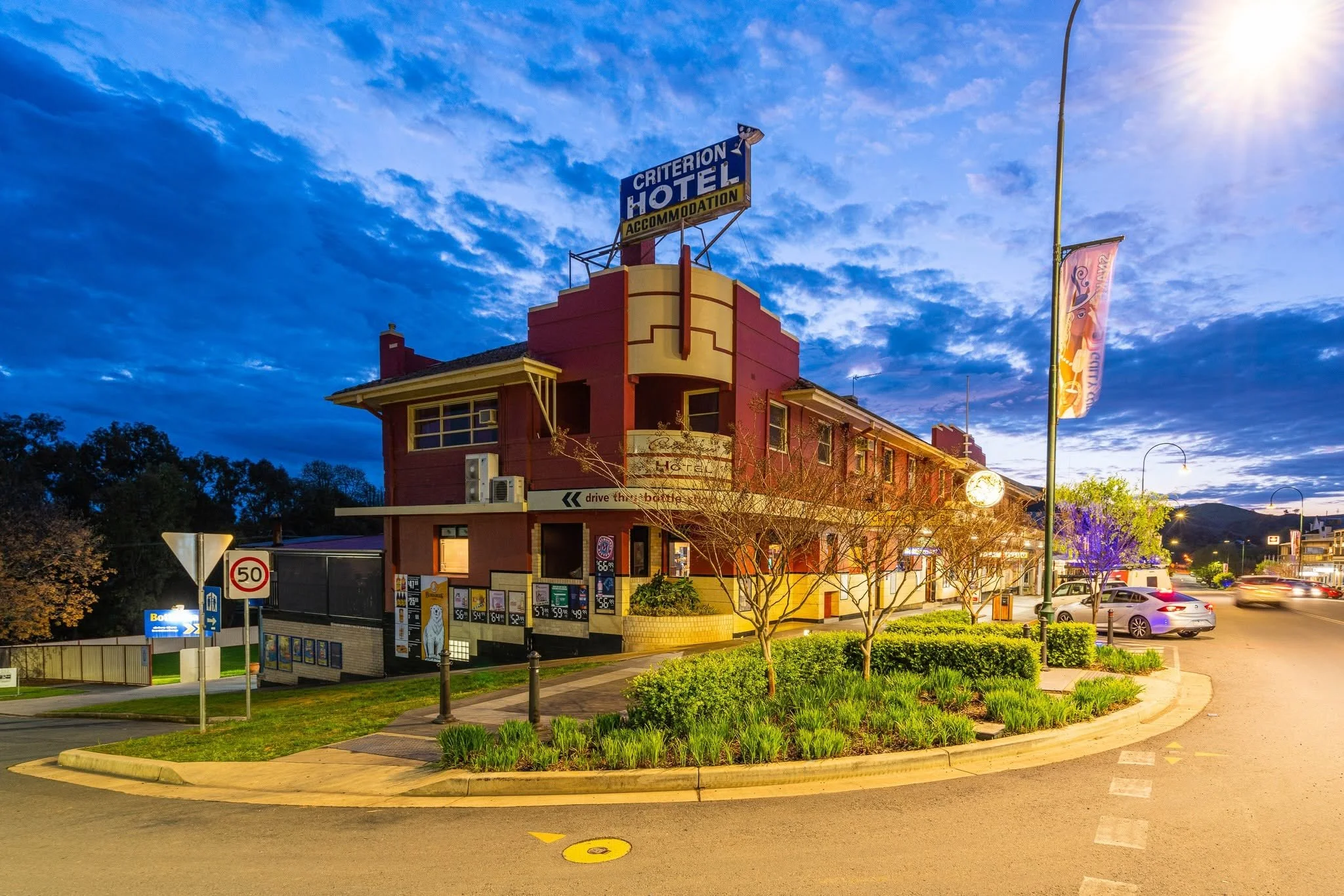 A two-story red and yellow building with a sign on the rooftop that says "Criterion Hotel" and "Accommodation." The building has a side balcony and windows, with streets and parked cars in front. The sky is partly cloudy with the sun shining from the