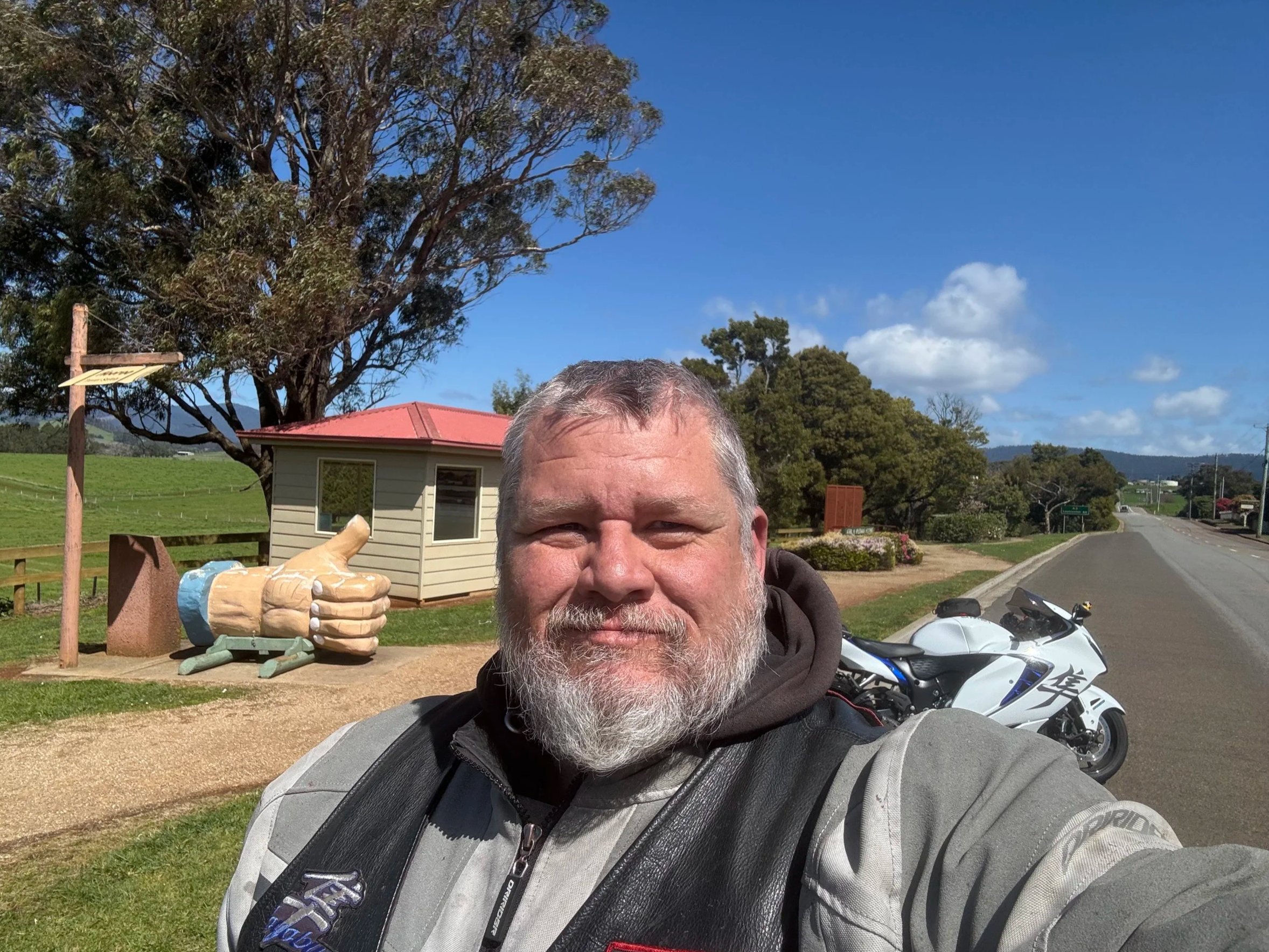 A man with gray hair and a beard taking a selfie outdoors on a sunny day. In the background, there is a sculpture of a large pair of hands giving a thumbs-up, a small building, and a landscape with trees, a road, and a clear blue sky with some clouds.