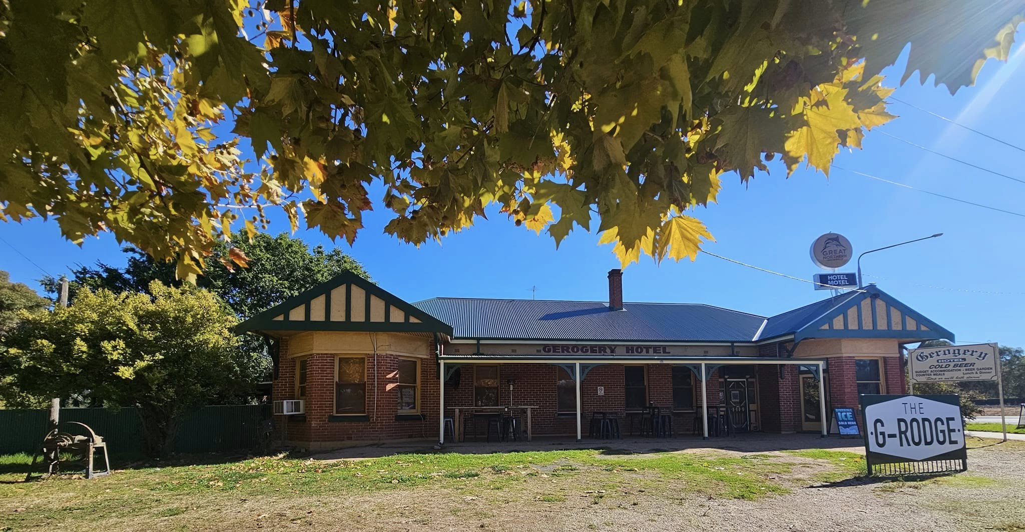 A brick building labeled 'Gerogery Hotel' with a blue metal roof, surrounded by trees with green and yellow leaves, and a sign for 'Berry's Corner' on the front lawn. The sky is clear and blue, with sunlight streaming from the upper right corner.