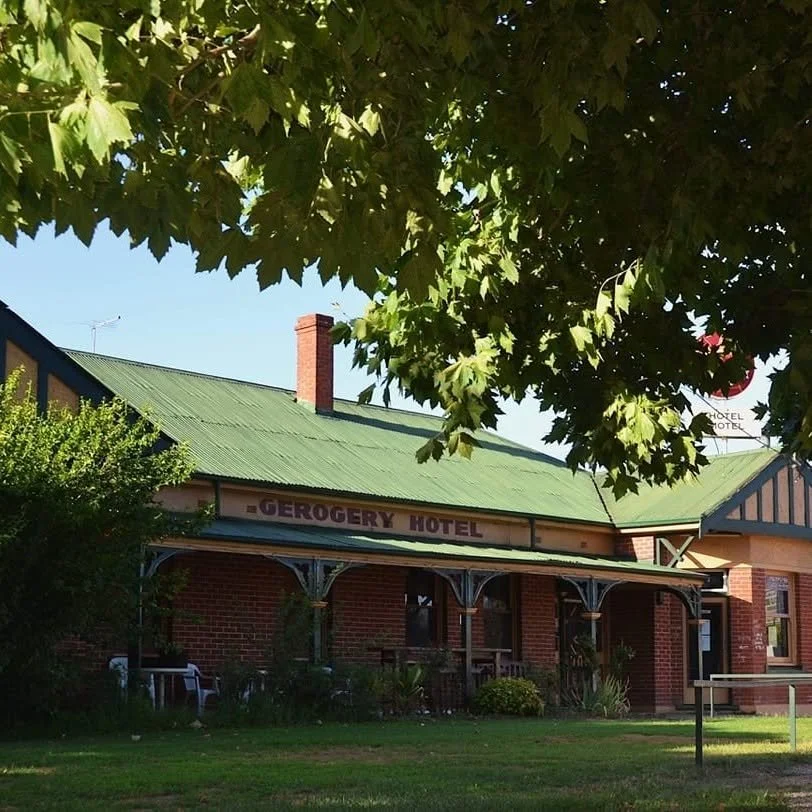 A brick building with a green metal roof, labeled 'Geregory Hotel'. There is a porch with decorative supports, some outdoor chairs, and trees providing shade.