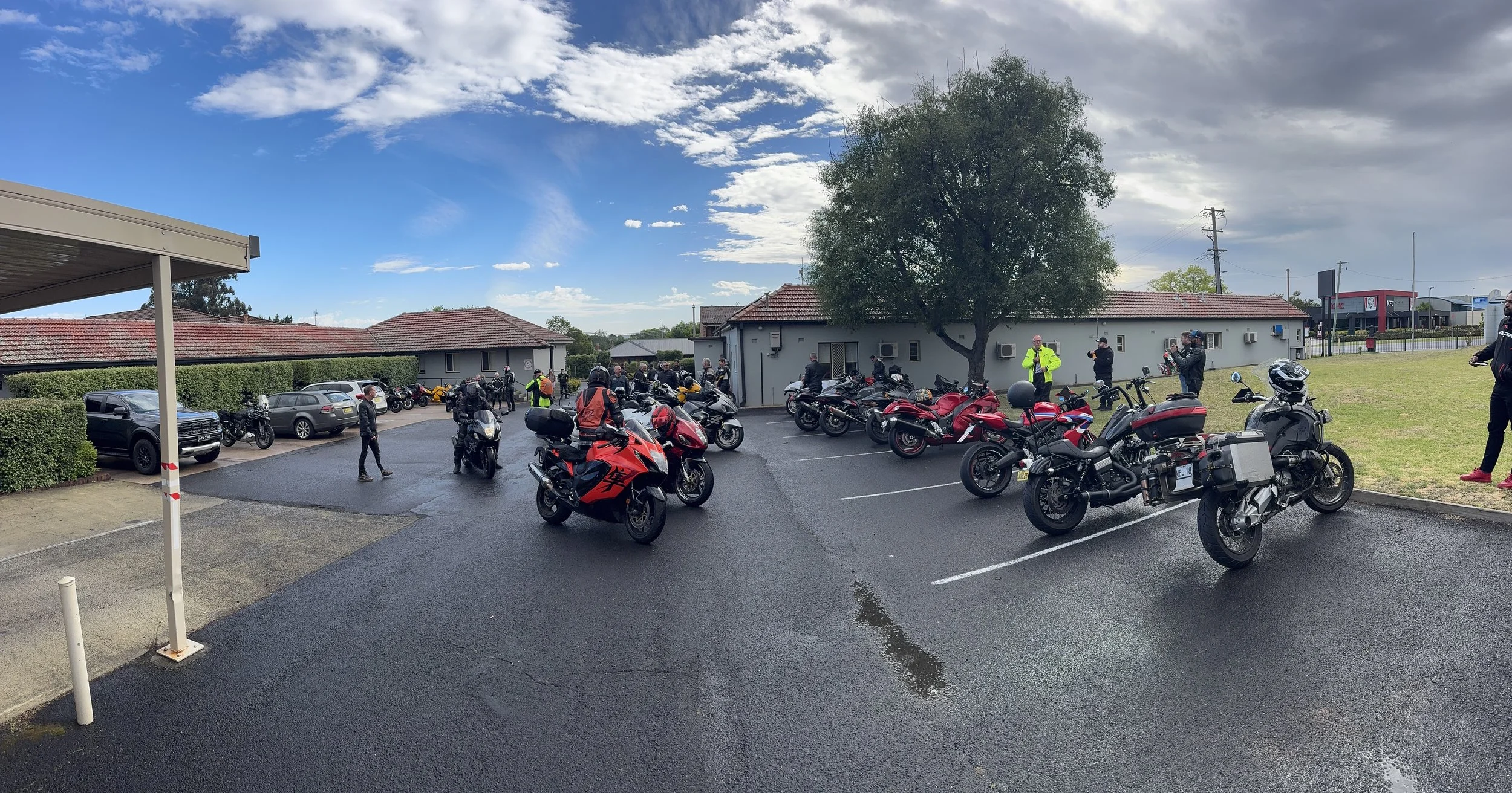 A parking lot with multiple motorcycles and a few cars, with people standing around, some wearing helmets. In the background, there are buildings, trees, and a partly cloudy sky.