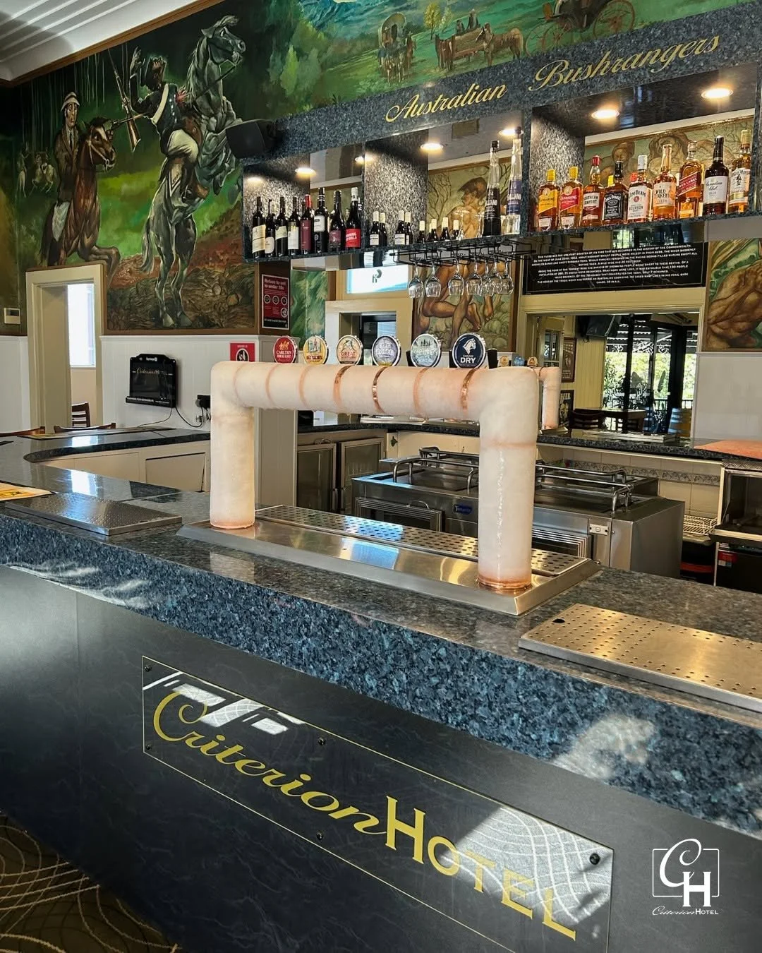 Interior of a bar or restaurant with a mural of Australian bushrangers and horses, bar shelves stocked with various liquor bottles, a tap connected to a large salt pipeline on the counter, and a sign with the text 'Citreion Hotel' on the front of the