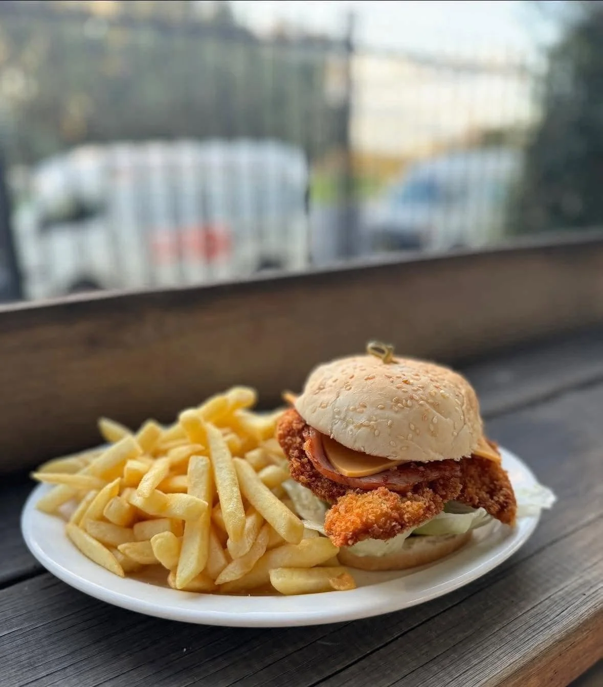 Plate with fried fish sandwich and French fries on dark wooden table, with blurred window view in the background.