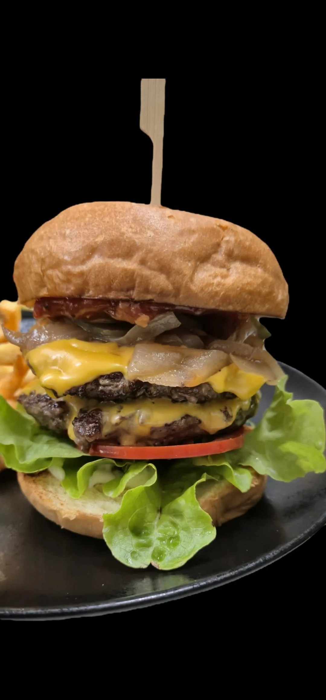 Close-up of a multi-layered cheeseburger with lettuce, tomato, beef patties, cheese, grilled onions, and a sandwich bun, held together with a wooden skewer, on a black plate against a black background.