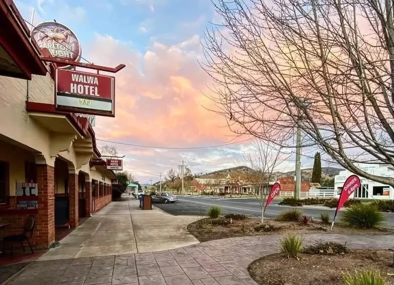 Street view of Walwa Hotel with signs indicating a bar, hotel, and dine-in options, along with a parking lot, sidewalk, trees, and a colorful sky at sunset.