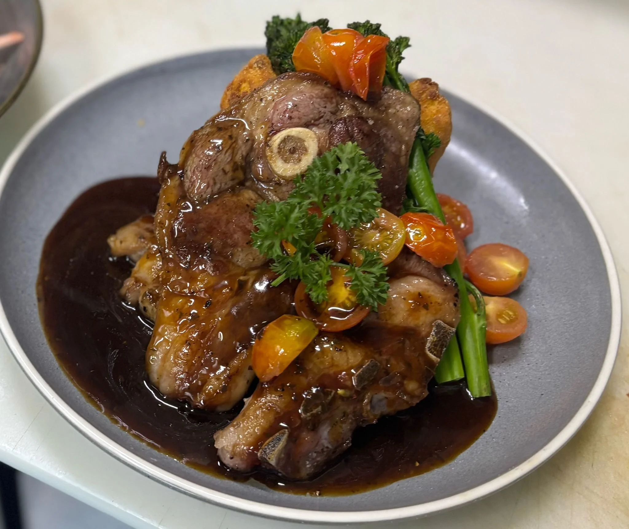 A plated dish featuring a meat chop topped with a rich brown sauce, garnished with fresh parsley and accompanied by roasted cherry tomatoes and green beans.