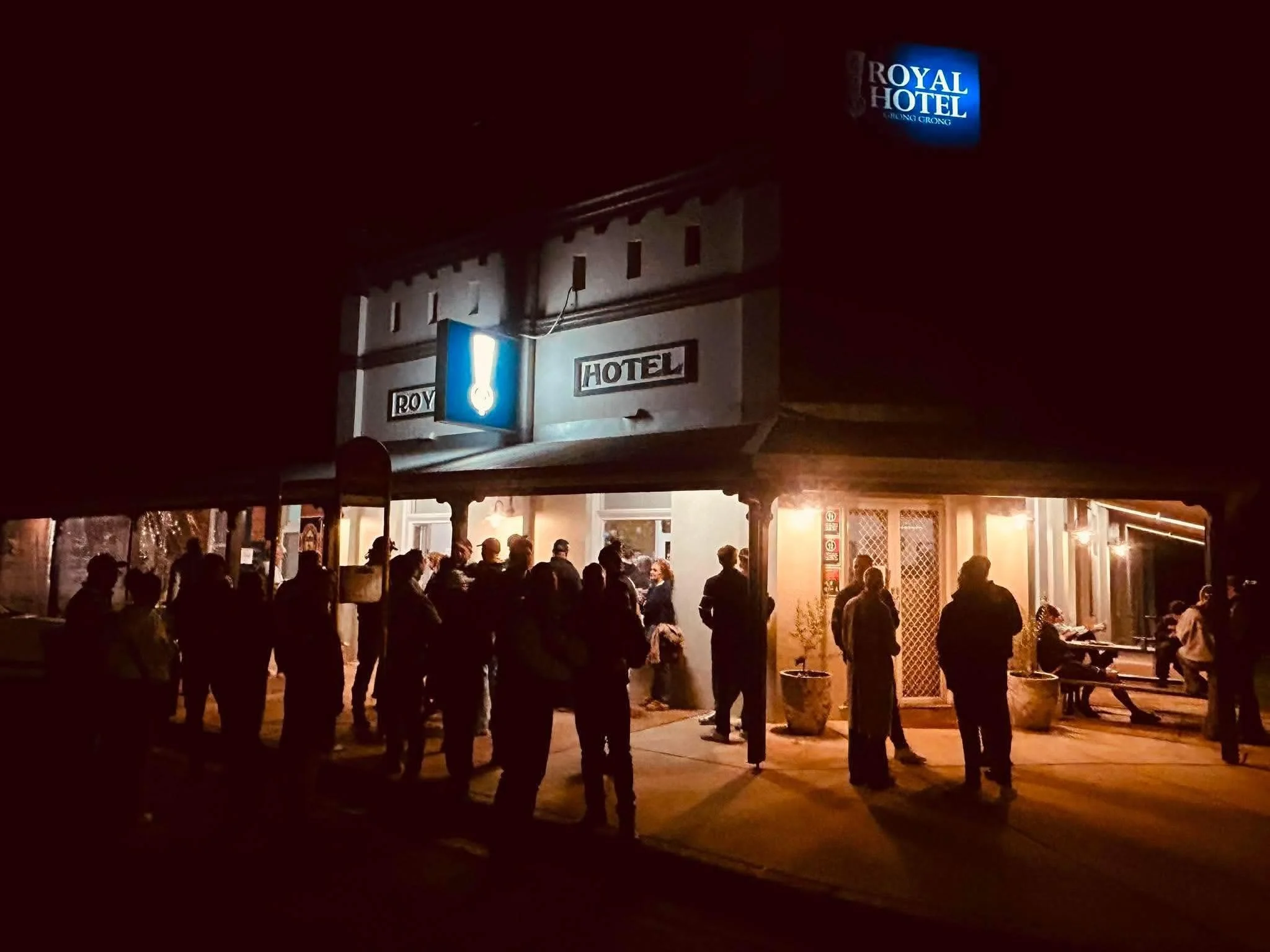 People standing outside a building at night, illuminated by warm lighting, with a sign that reads "Royal Hotel" and a bright blue sign above it.