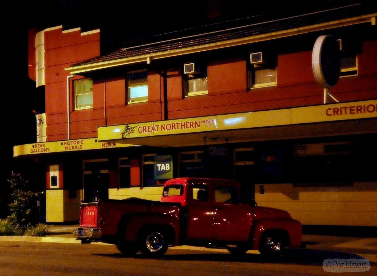 Nighttime street scene featuring a vintage red pickup truck parked in front of a building with yellow signage for Great Northern Brewing Co. The building has windows with air conditioning units and a mural advertising rear balcony, historic murals, m