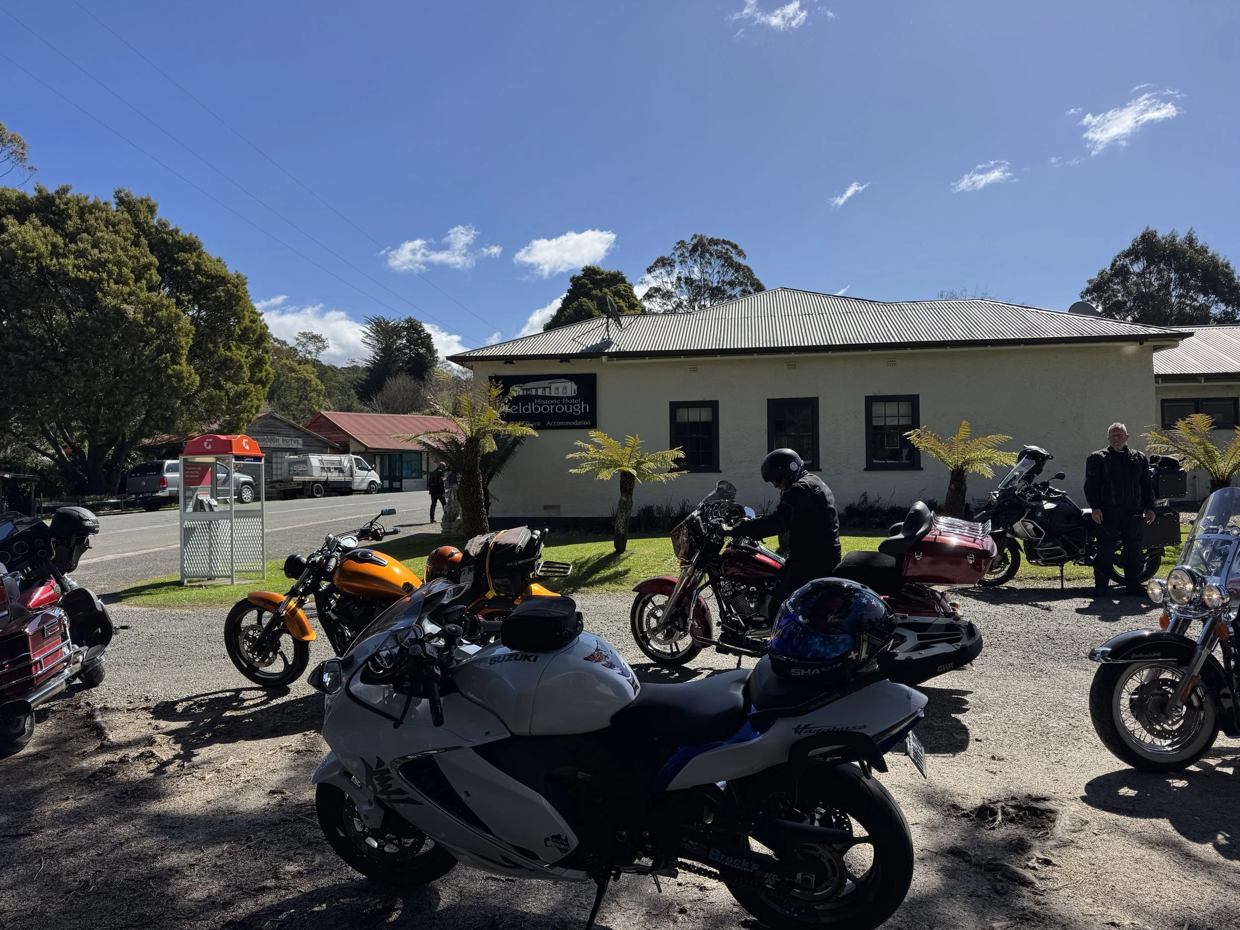Motorcycles parked outdoors in front of a white building with trees and a blue sky with clouds.