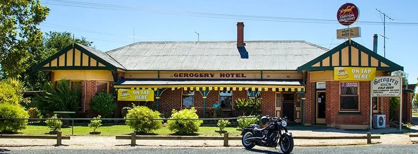 Front view of Gerogery Hotel, a single-story brick building with a metal roof and green trim, yellow signs advertising cold beer on tap, and a motorcycle parked outside.