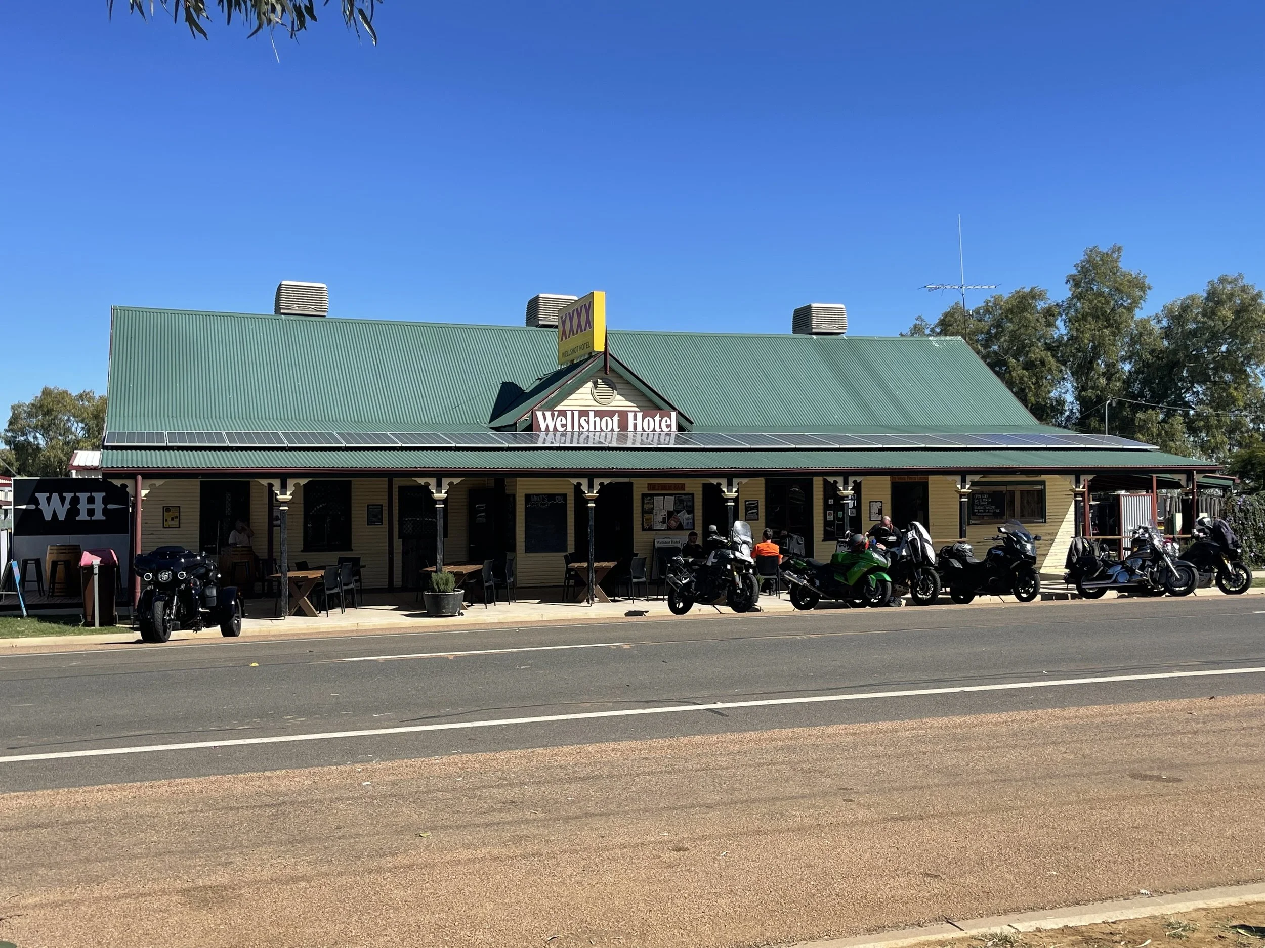 A building with a green metal roof, labeled as Wellshot Hotel, with motorcycles parked in front and trees on the side. The sky is clear and blue.