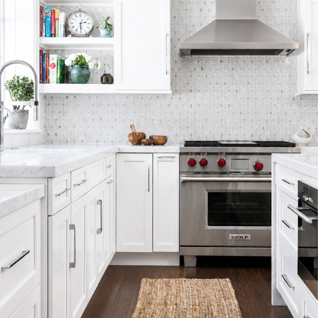 Modern kitchen with white cabinets, marble countertops, a stainless steel Wolf oven with red knobs, a range hood, open shelving with books and decorative items, a window with a plant, and a woven rug on a dark wood floor.