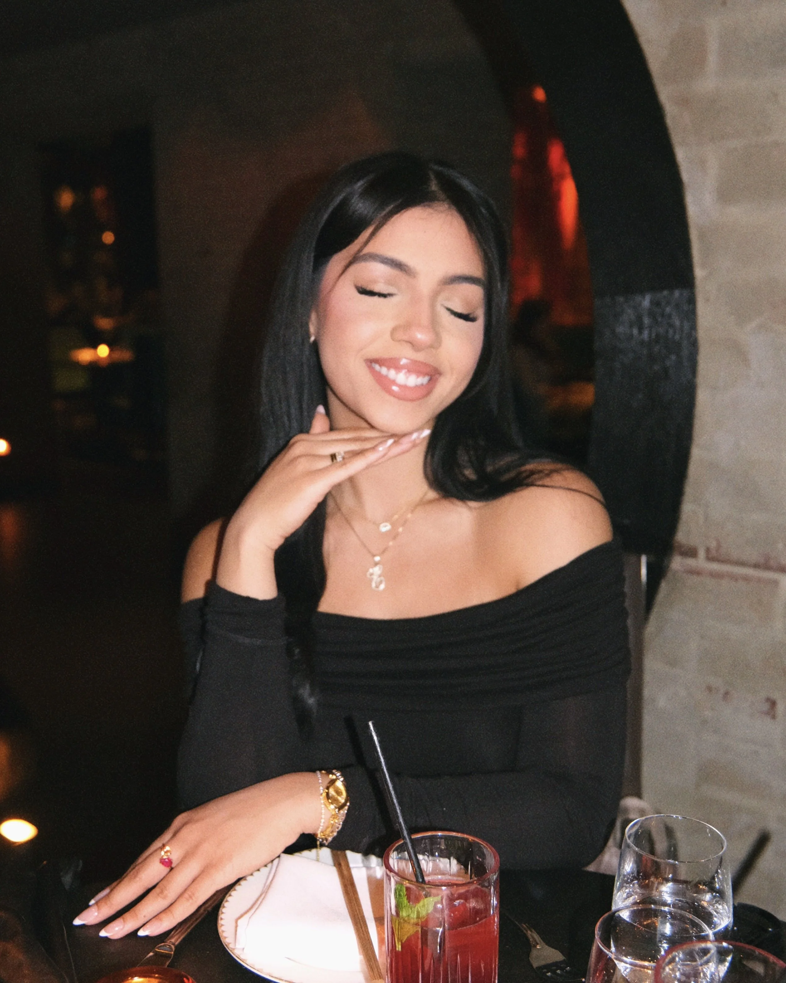 A woman with long dark hair, wearing a black off-shoulder top, jewelry, and makeup, sitting at a restaurant table with drinks, embracing her hands near her chin with closed eyes and smiling, in a dimly lit setting.