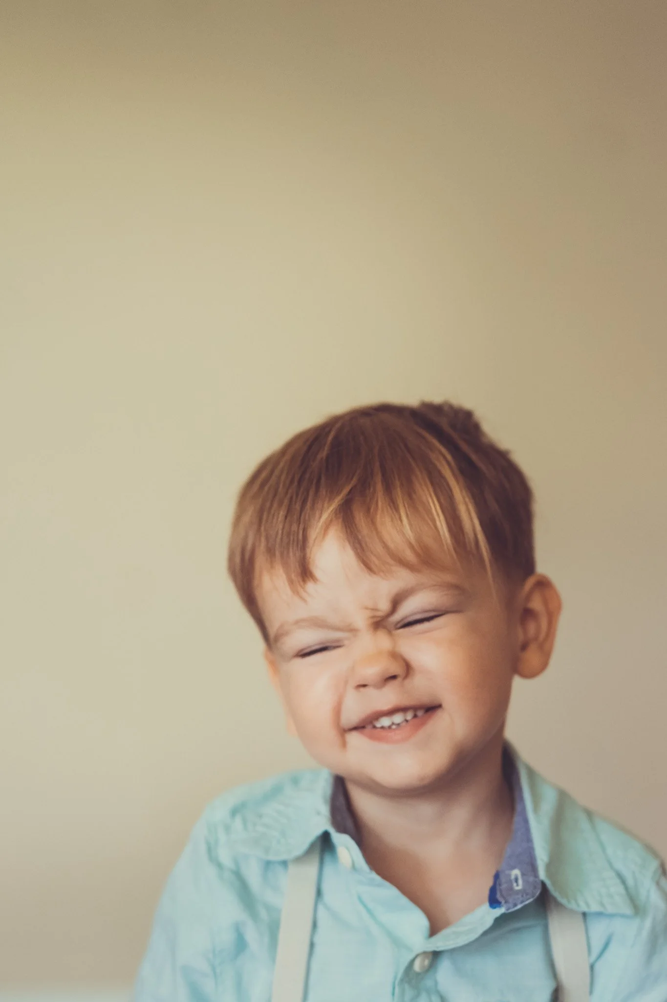 A young boy with red hair and a big smile, showing his teeth, wearing a light blue shirt and suspenders, with a plain beige background.