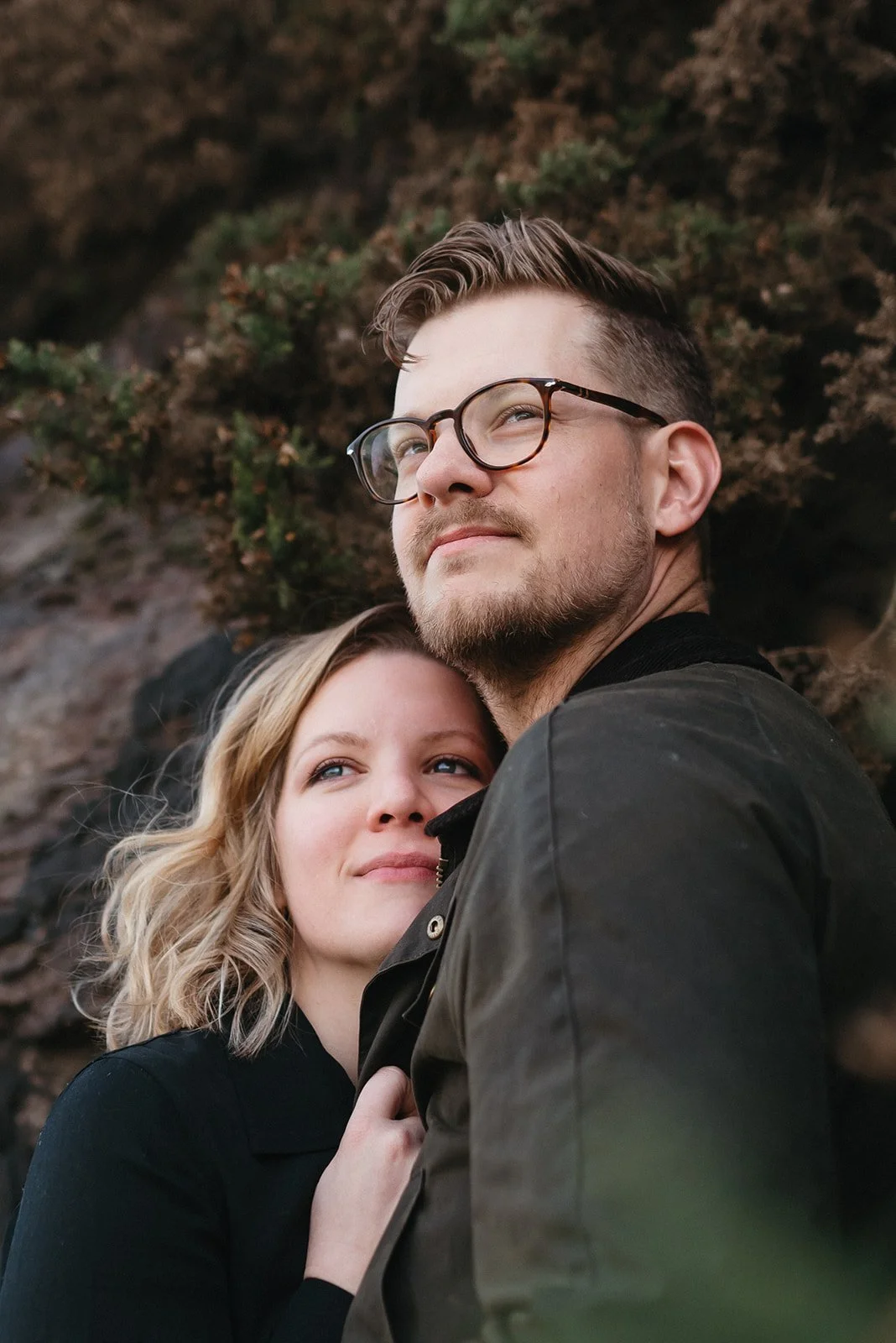 A man and woman close together outdoors, with the woman resting her head on the man's chest, both smiling softly.