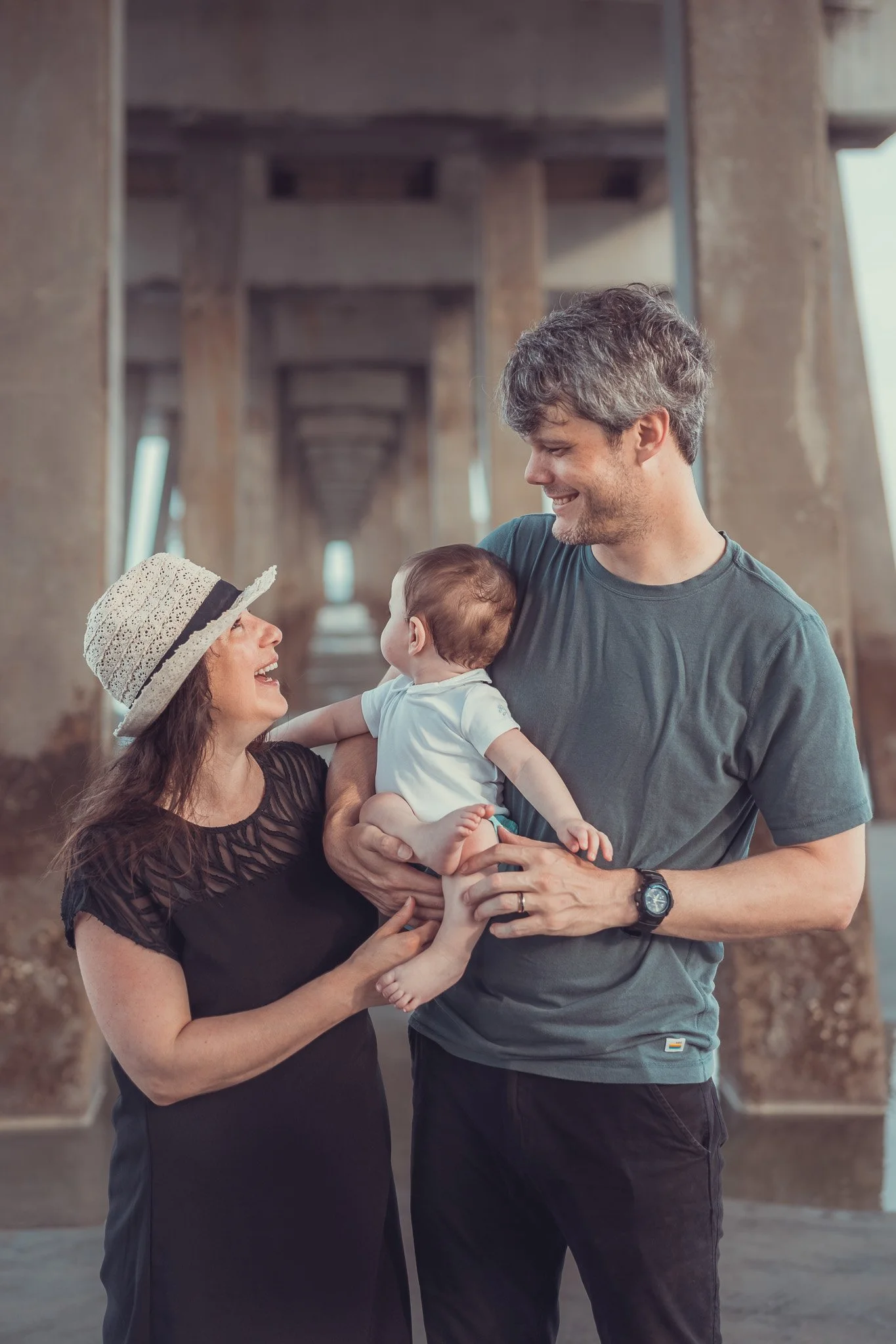 A woman wearing a straw hat and a black dress holding a baby, smiling while looking at a man wearing a gray t-shirt and black pants, standing on a bridge with concrete pillars in the background.