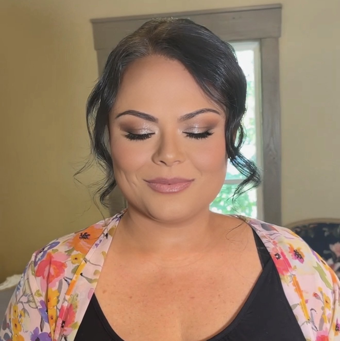 Close-up of a woman with short dark hair and makeup, wearing a black top and a colorful floral robe, smiling softly with eyes closed indoors near a window.