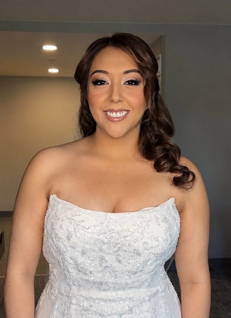 A woman in a white, strapless wedding dress with intricate lace and beading, smiling in soft glam makeup