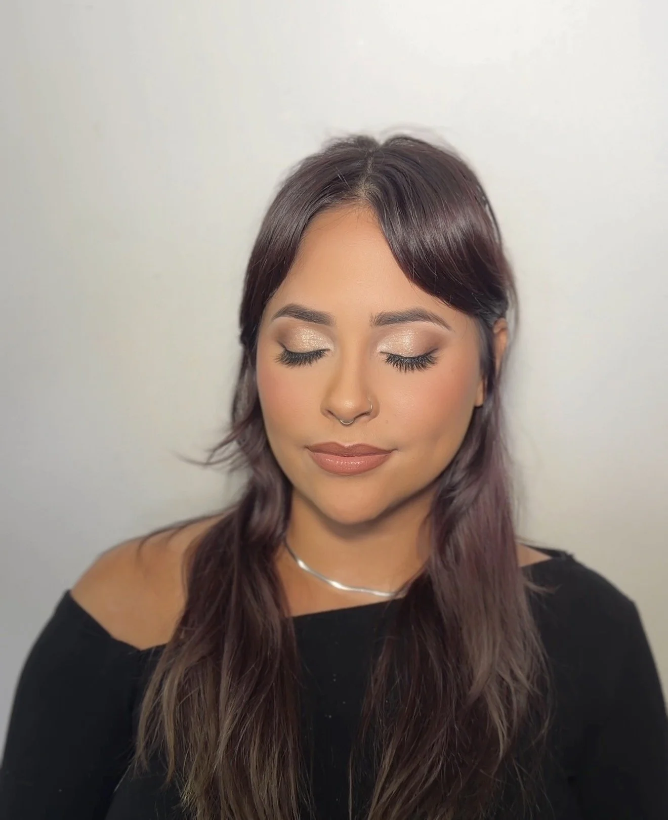 A woman with long, wavy brown hair and light makeup, including gold eyeshadow, a nose ring, and glossy lips, is smiling softly with her eyes closed against a plain white background. She is wearing a black off-the-shoulder top and a silver necklace.