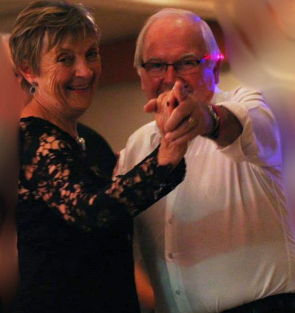 An elderly man and woman smiling and dancing together at a social event, with the man pointing towards the camera.