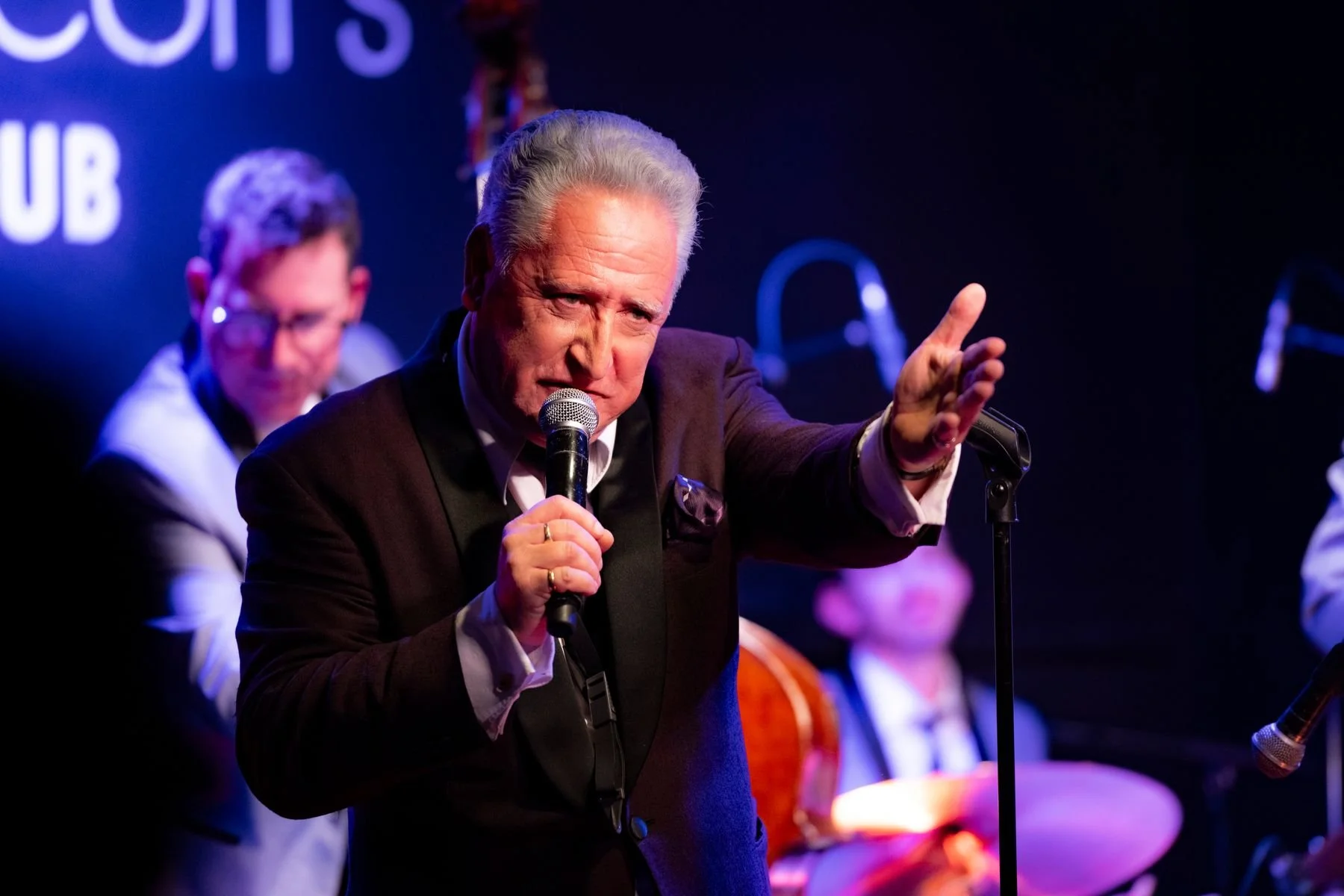 An older man with gray hair and a serious expression is speaking into a microphone on a stage, gesturing with his right hand. Behind him, there is a man with glasses and dark hair, and a person in a traditional Mexican dress, indicating a cultural or musical event.