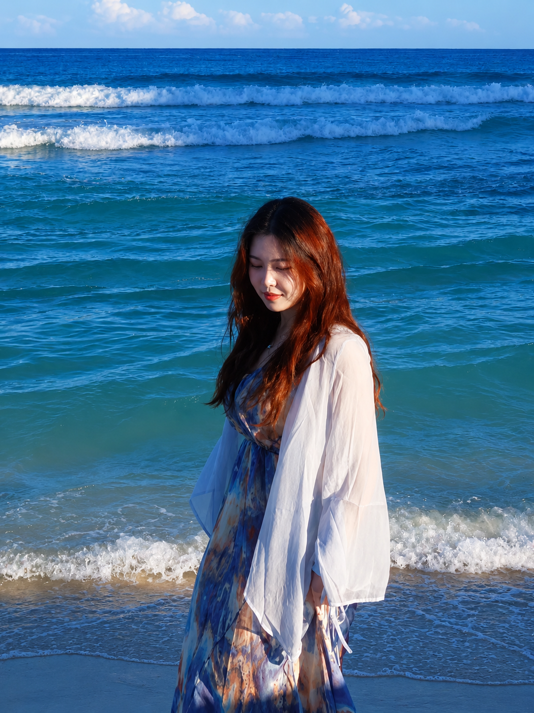A woman with long red hair wearing a flowing dress and a white cover-up standing on the sandy shore at the beach with ocean waves in the background.