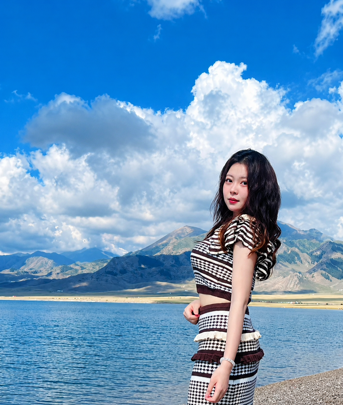 Young woman with long dark wavy hair standing near a lake with mountains and a blue sky with clouds in the background.