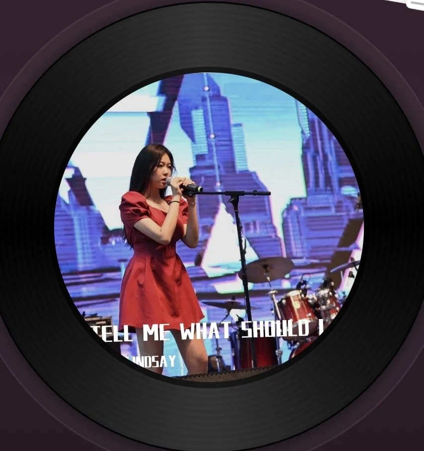 Young woman in red dress singing into a microphone on stage, with musical instruments in the background and a cityscape display on a large screen behind her.