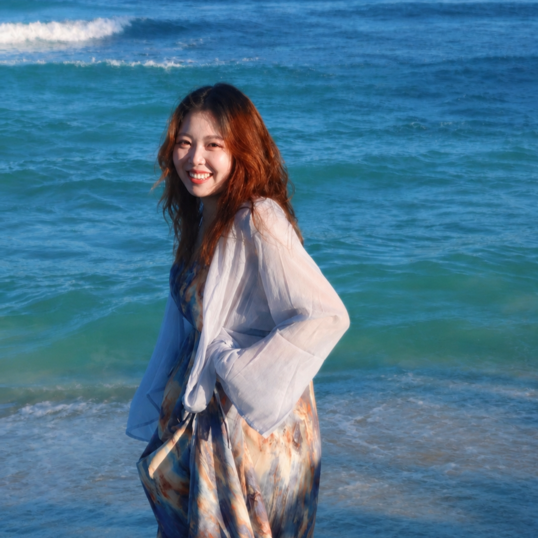 A woman with wavy brown hair smiling, standing by the ocean with waves behind her.