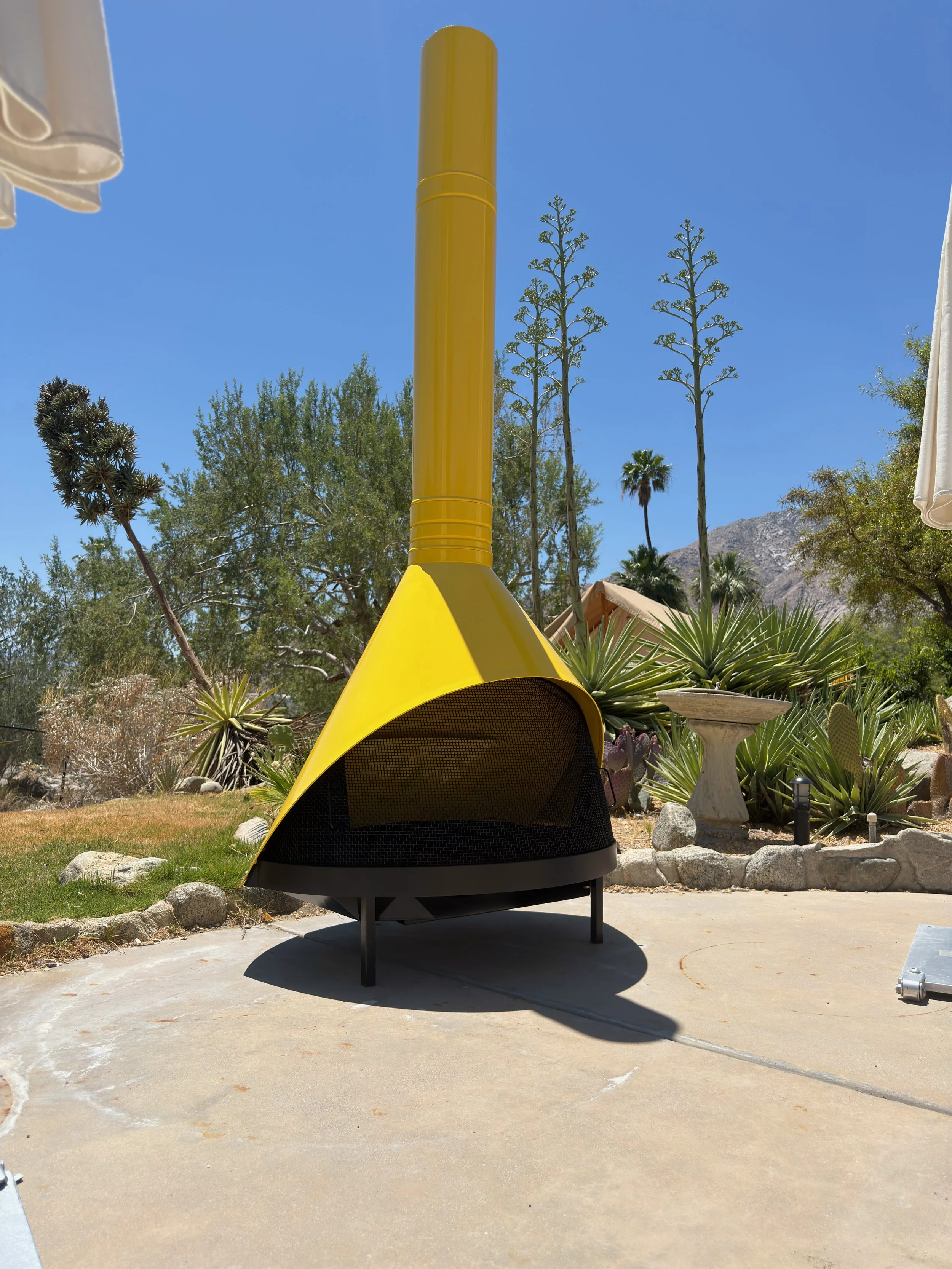 A yellow Cornhole game set up outdoors on a concrete patio, with desert plants, trees, and a mountain in the background on a sunny day.