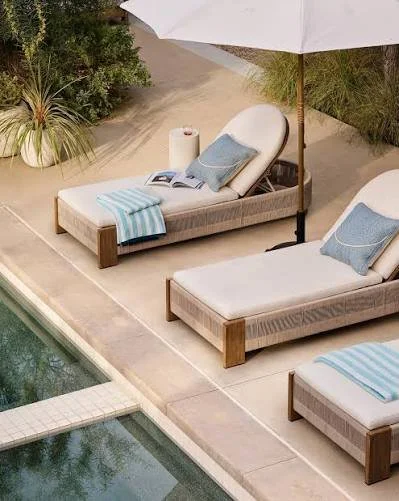 Two lounge chairs with cushions and towels next to a swimming pool, with an umbrella providing shade, and potted plants nearby.