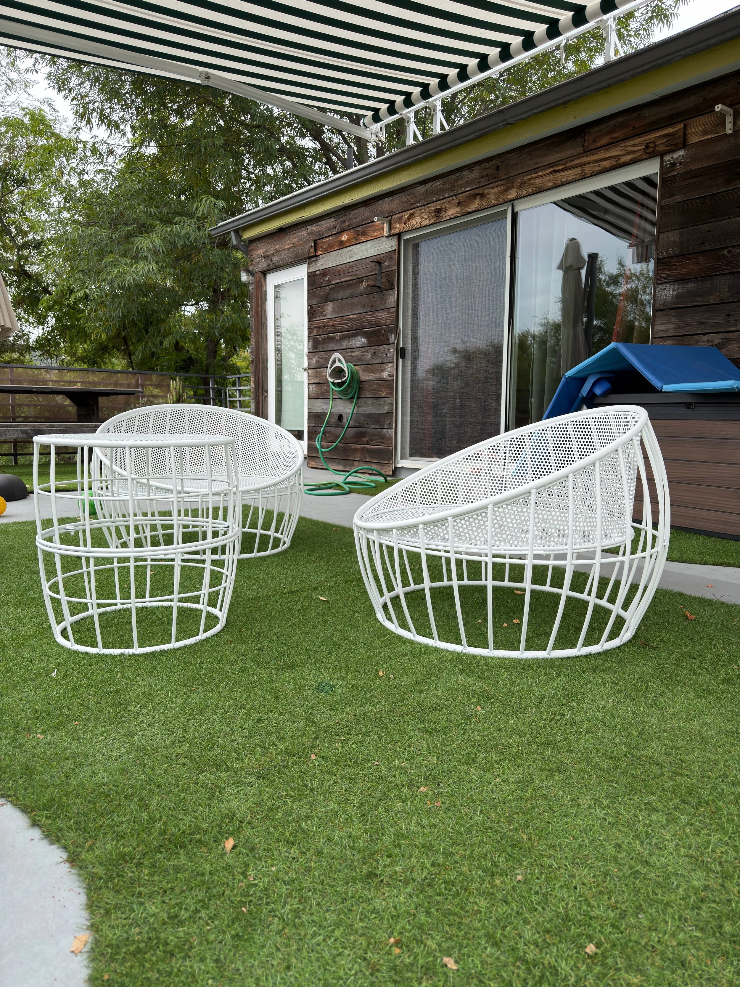 A backyard patio with white metal outdoor chairs, a wrapped hot tub, a green garden hose on the house, a partial view of a sliding glass door with a screen, and a green lawn with a concrete walkway, surrounded by a black metal fence and trees.