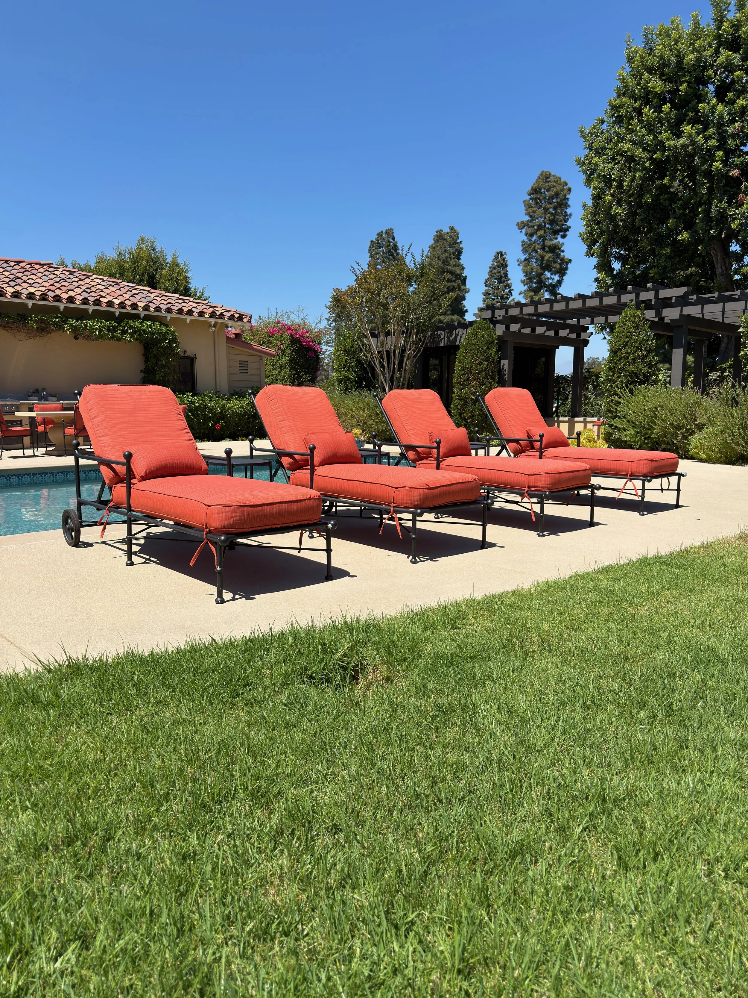 Loungers with red cushions arranged by the poolside on a sunny day, with a house, trees, and a clear blue sky in the background.