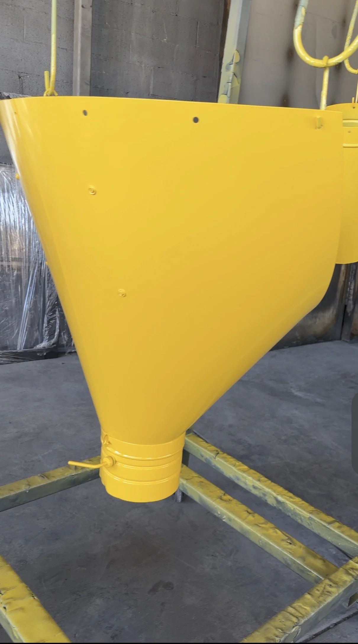 Yellow industrial funnel connected to a pipe, standing on a metal frame in a warehouse or factory setting.