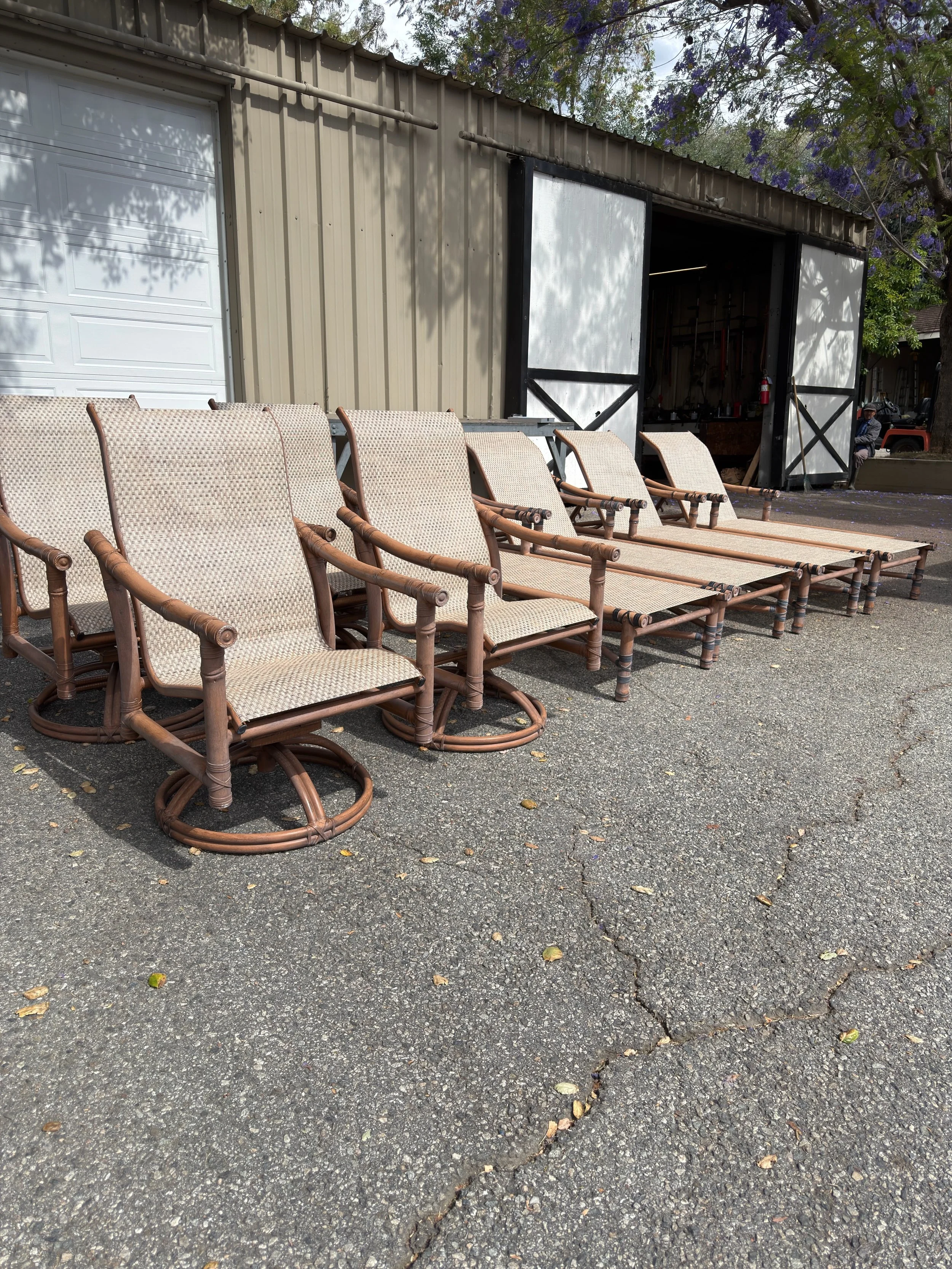 Six beige outdoor chairs with wooden armrests arranged in a row outside a metal building with a large garage door and sliding doors. There are trees with purple flowers overhead and a person sitting in the background.