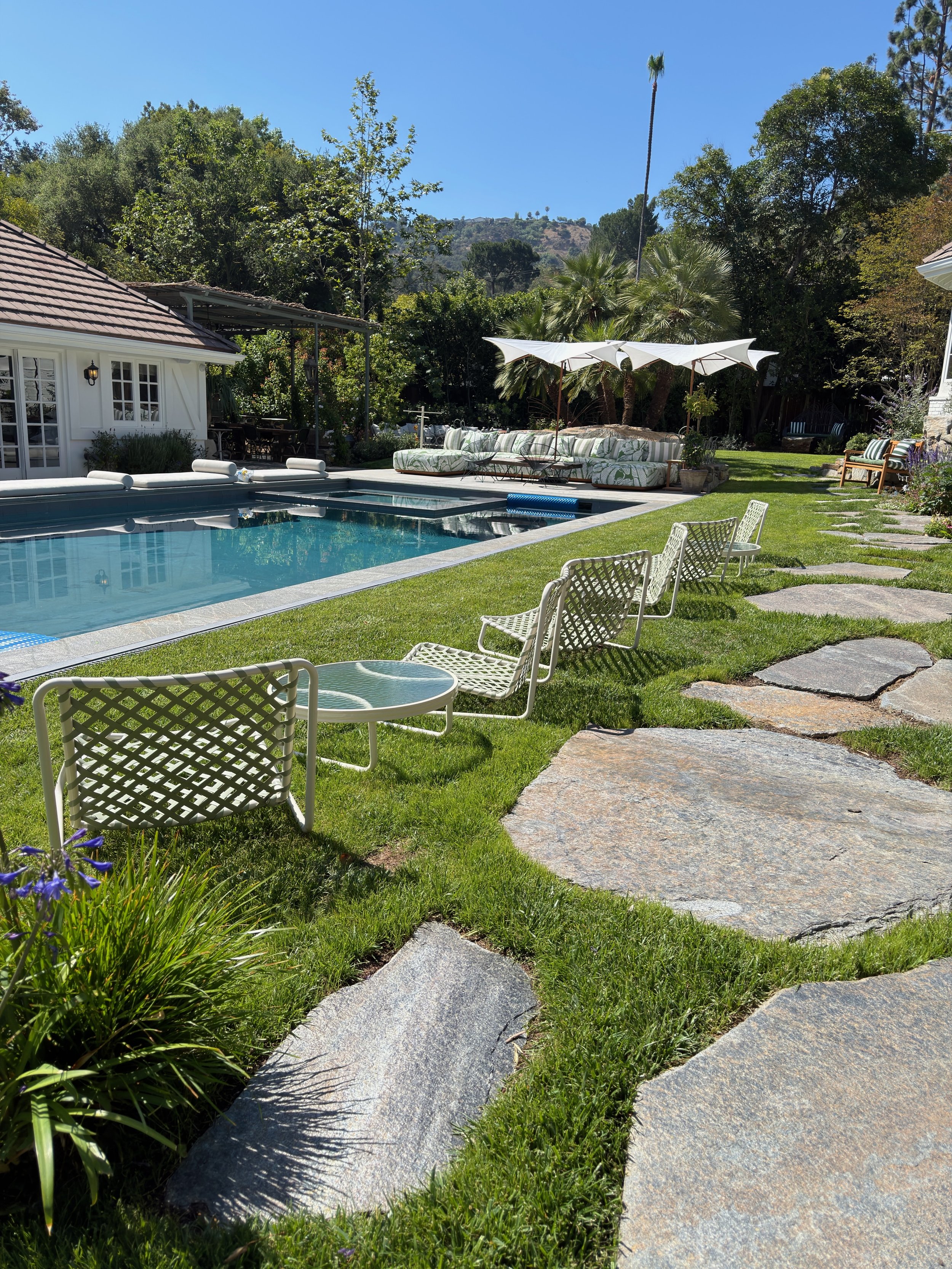 A backyard with a swimming pool, outdoor furniture including lounge chairs, a patio, lush green grass, trees, and hills in the background under a clear blue sky.