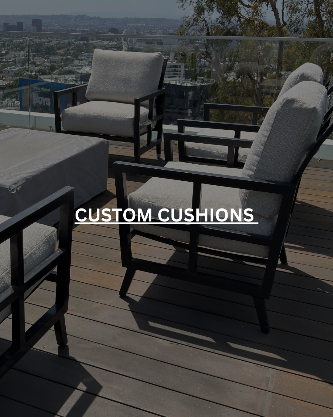 Outdoor rooftop patio with wooden flooring, several gray cushioned chairs with black frames, and a cityscape view in the background. Text overlay reads 'Custom Cushions'.