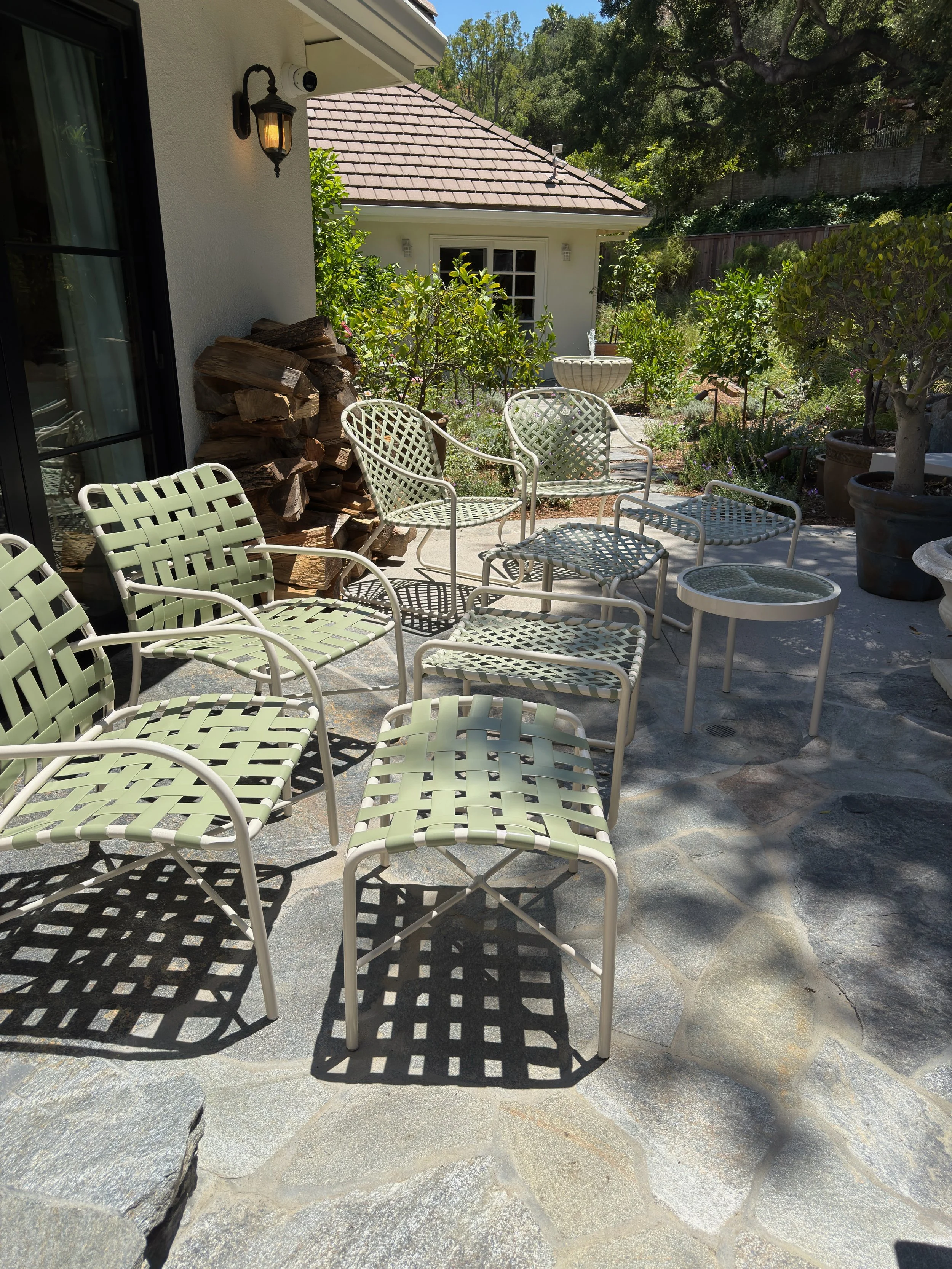 A patio area with multiple white metal chairs with green woven seats and backs, a small round glass-top table, potted plants, a woodpile, a fountain, and a garden with greenery and trees in the background.