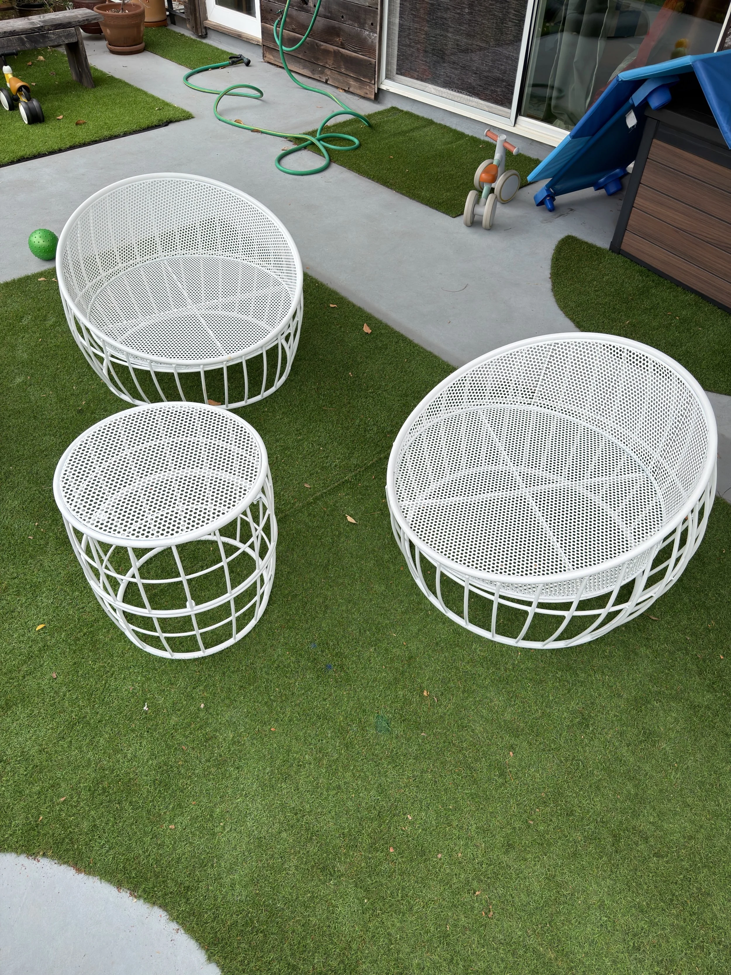 A backyard patio with two white wireframe circular chairs on grass, a small green rubber ball nearby, a concrete walkway, a garden hose, children's tricycle, a small blue and orange plastic playhouse, and a wooden outdoor storage shed.